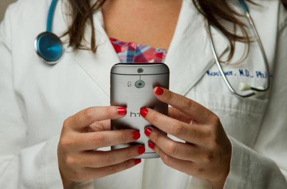 A doctor in a white coat with a stethoscope holds a smartphone, showcasing red-painted nails against a plaid shirt background.